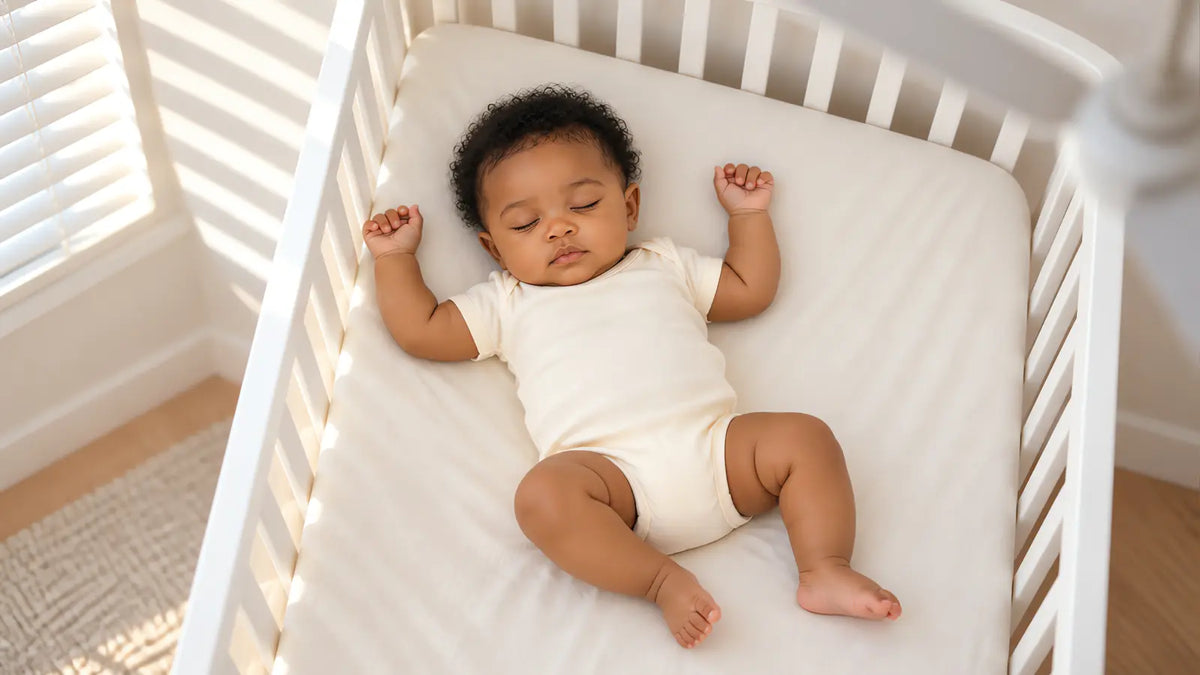 A baby asleep in a single thin layer, with the afternoon light coming through the shutters beside the crib.