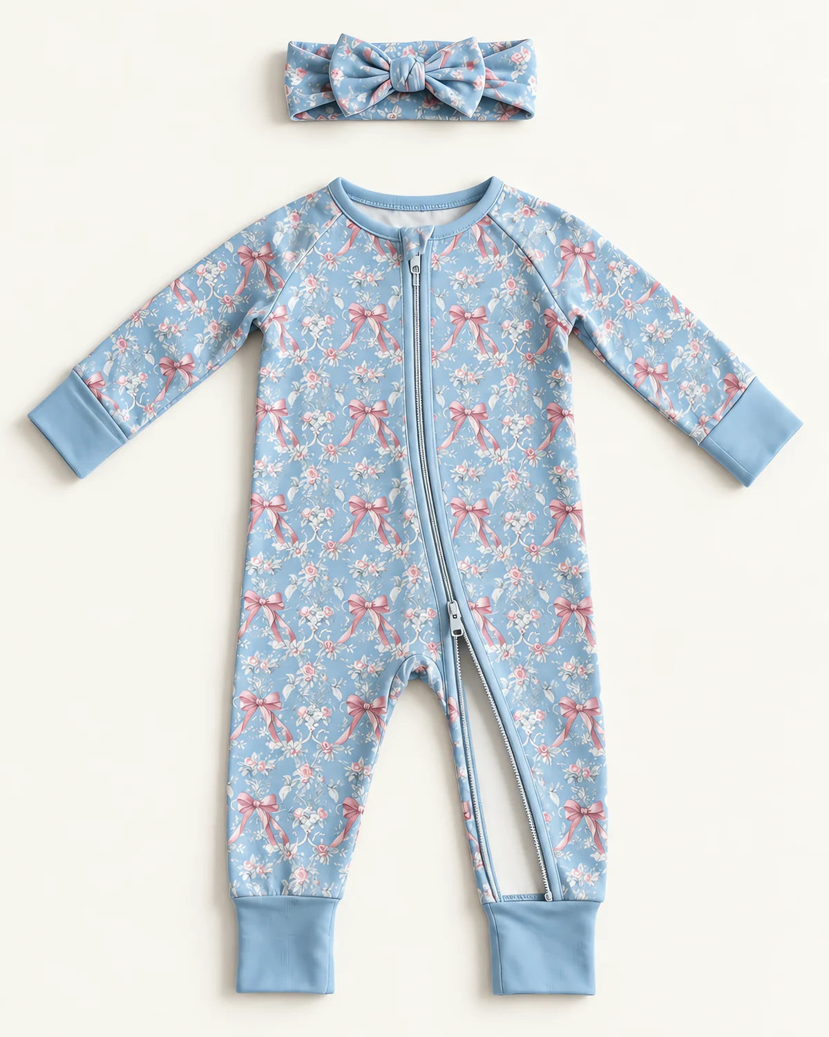 Flat lay of the JOWI Blue Rose Toile set on a plain pale background: the zip romper laid out open, unzipped from the ankle up so the teeth and the slider show, and the matching bow headband in the same rose and ribbon print lying separately above it