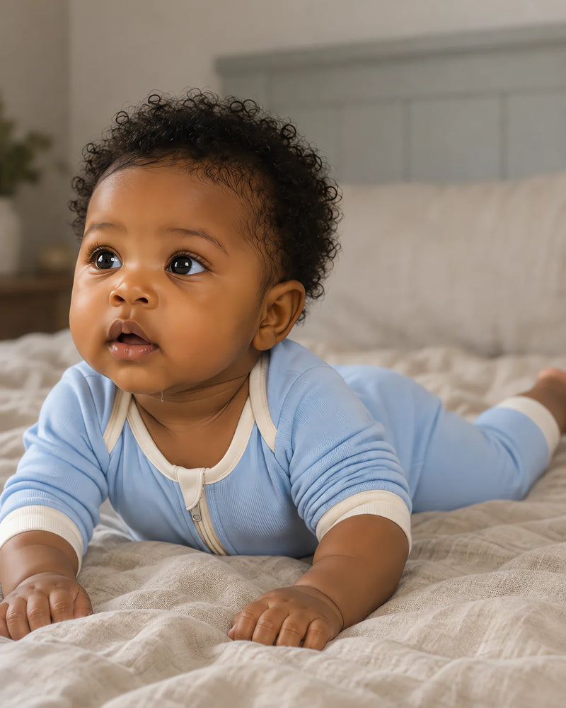 Six-month-old baby during tummy time on a crumpled linen sheet, wearing the JOWI plain light blue zip romper with cream cuffs and cream neck binding