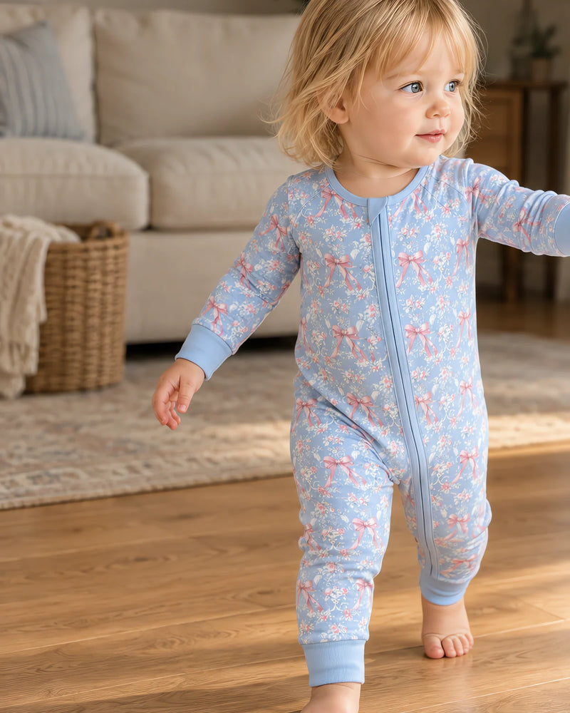 Toddler taking a step barefoot on a wooden floor, wearing the JOWI zip romper in light blue with a rose and pink bow print