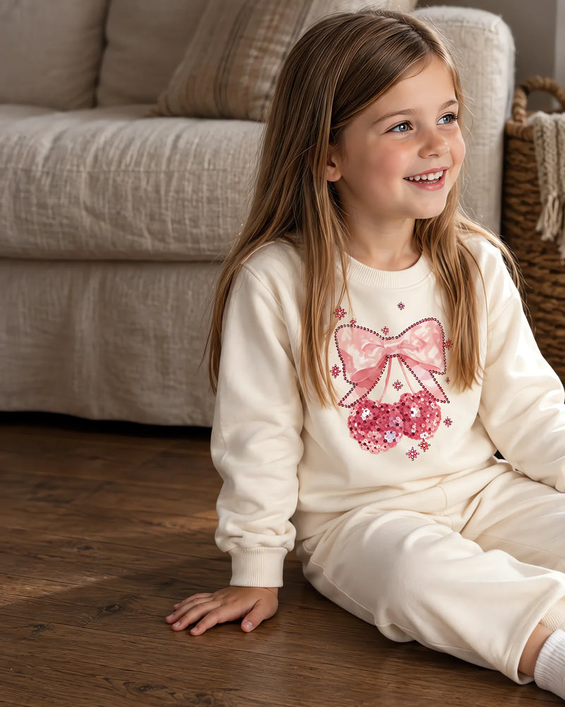 Five-year-old girl sitting on the floorboards of a living room wearing the JOWI cream sweatshirt and jogger set, with the pink bow and the sequin cherries on the chest