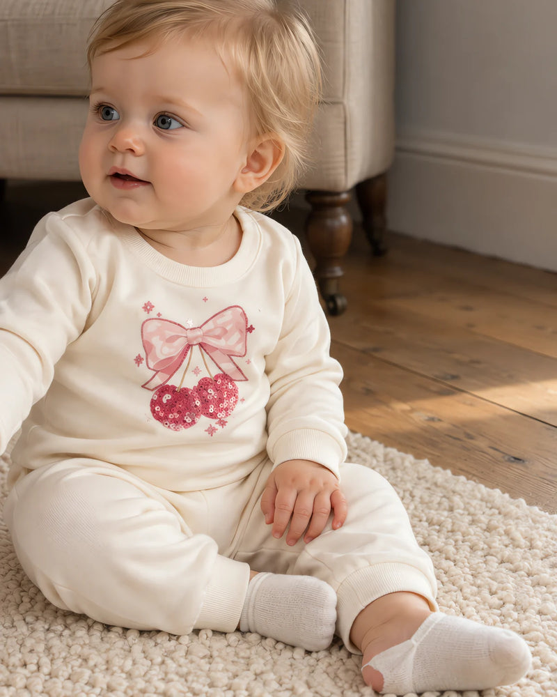 Baby sitting on a cream rug on a wooden floor, wearing the JOWI cream set with a pink bow and cherries on the chest and plain sleeves